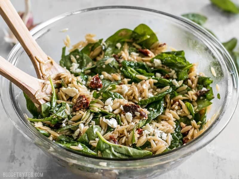 Side view of a glass bowl full of Spinach and Orzo Salad with wooden salad tongs