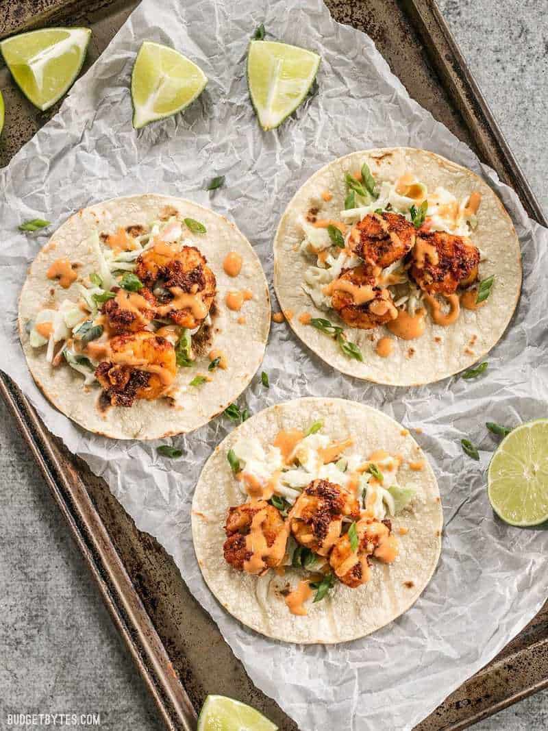 Three open Blackened Shrimp Tacos on a metal baking sheet with lime wedges nearby