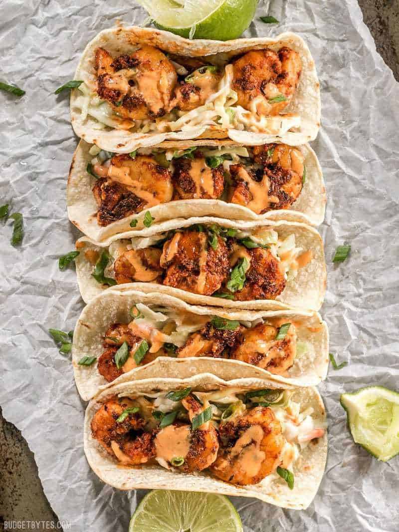 Five Blackened Shrimp Tacos lined up on parchment paper with limes all around