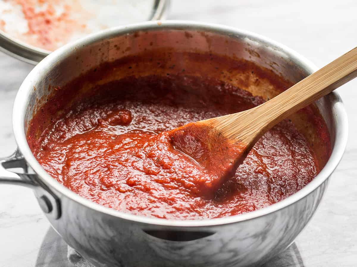 Side view of a pot full of pizza sauce with a wooden spoon in the center.