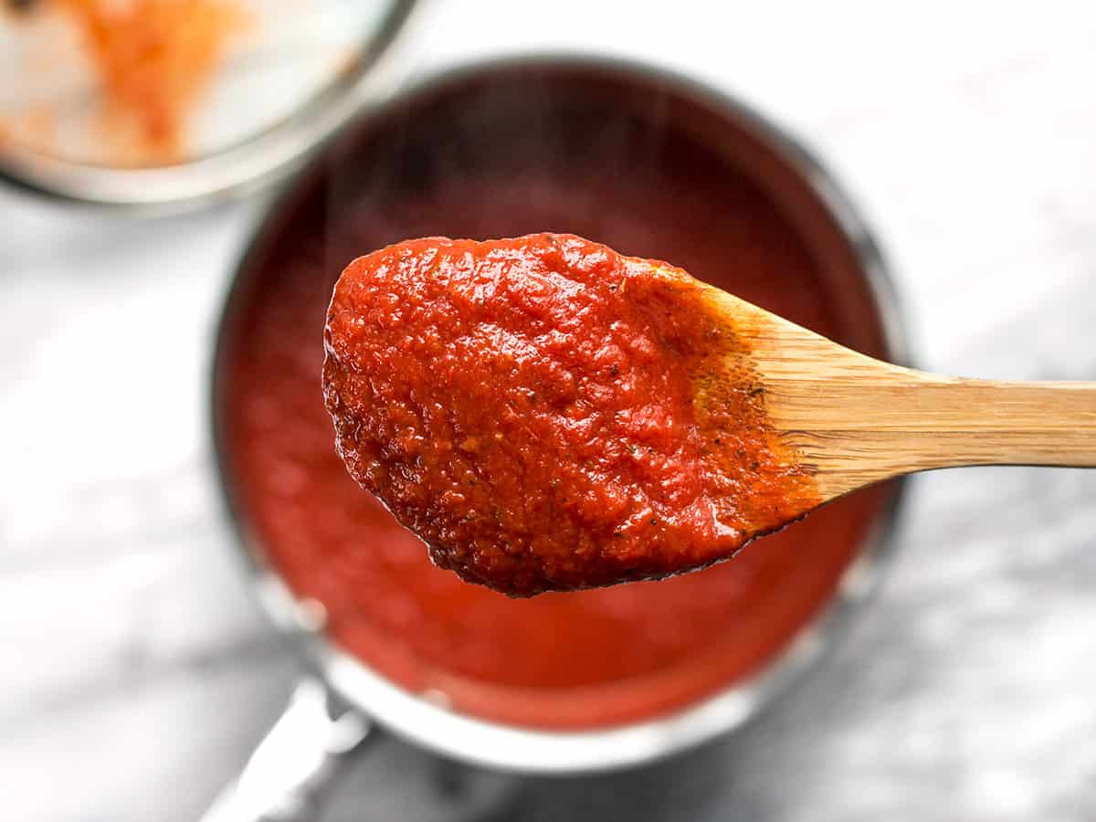 Close up of a wooden spoon full of pizza sauce held over a sauce pot.