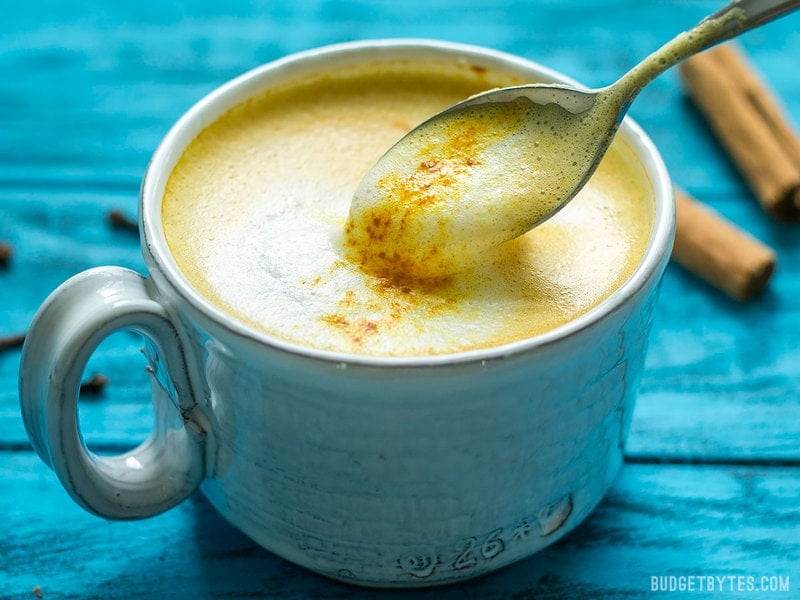 Front view of a mug of Golden Chai with a spoon lifting the foamy milk on top