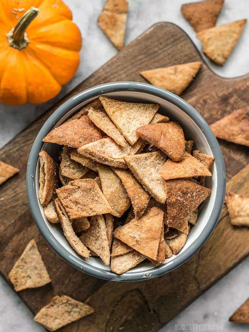 A bowl of Cinnamon Pita Crisps on a wooden cutting board next to a pumpkin