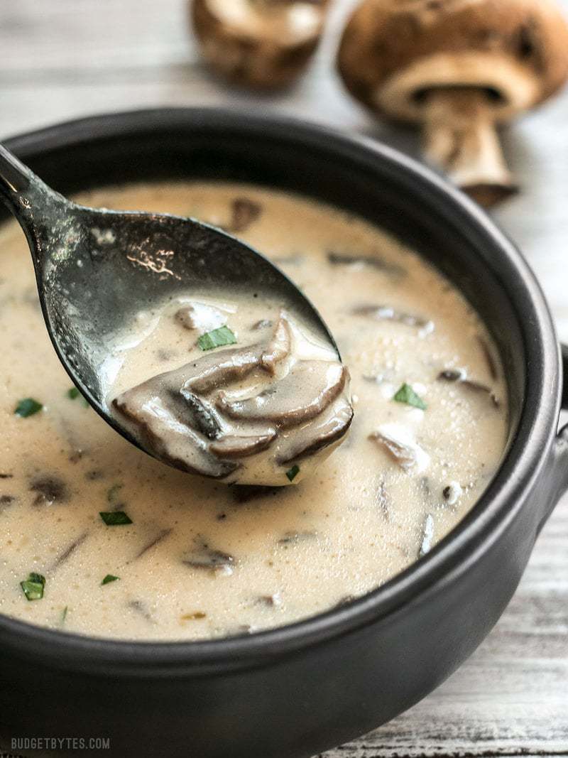 Close up of a spoonful of Creamy Garlic Mushrooms Soup in a black bowl with whole mushrooms in the background
