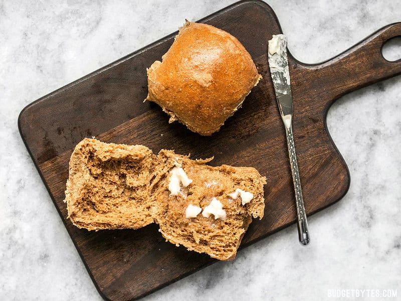 Two Sweet Molasses Dinner Rolls on a wooden cutting board, with butter smeared on top
