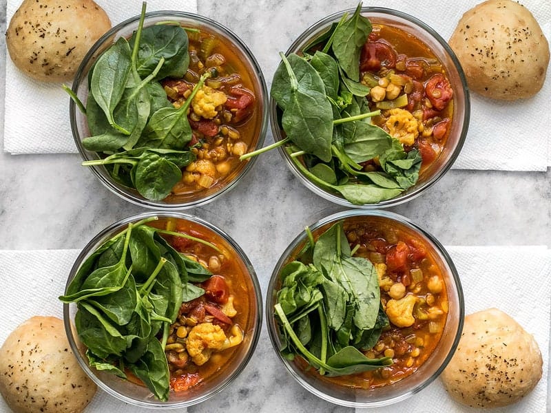Four Moroccan Lentil and Vegetable Stew meal prep bowls with spinach and focaccia rolls on the side