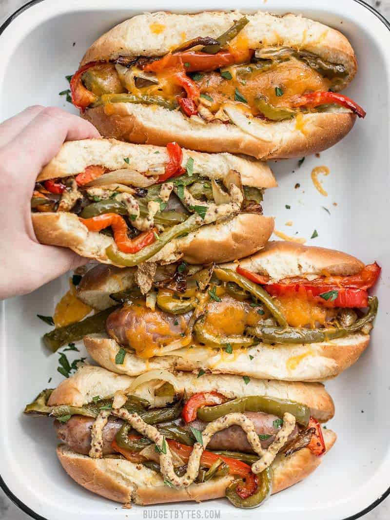 A hand taking one of the Roasted Bratwurst with Peppers and Onions in a bun with melted cheese and mustard.