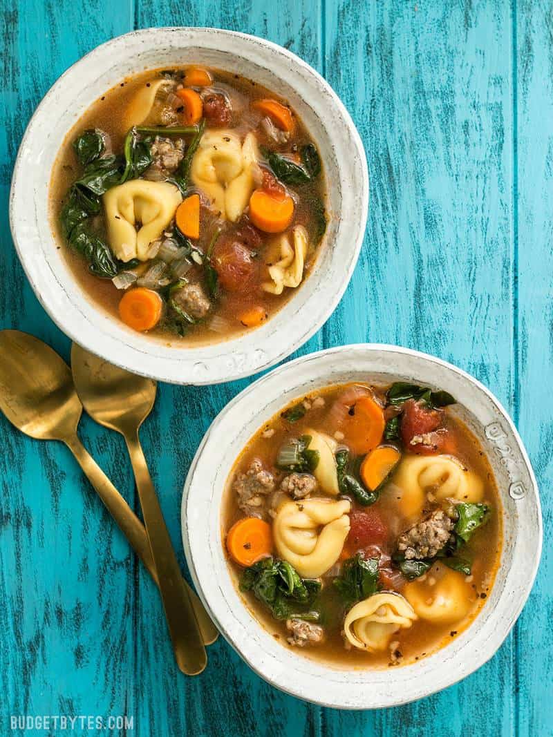 Two bowls of Sausage and Tortelloni Soup on a blue background with gold spoons