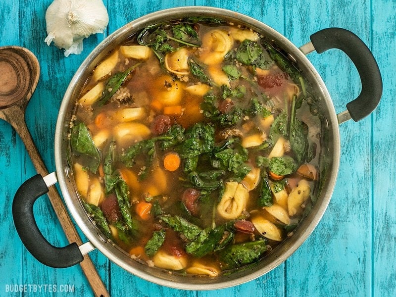 A big pot of Sausage and Tortelloni soup with floating spinach and a wooden spoon on the side