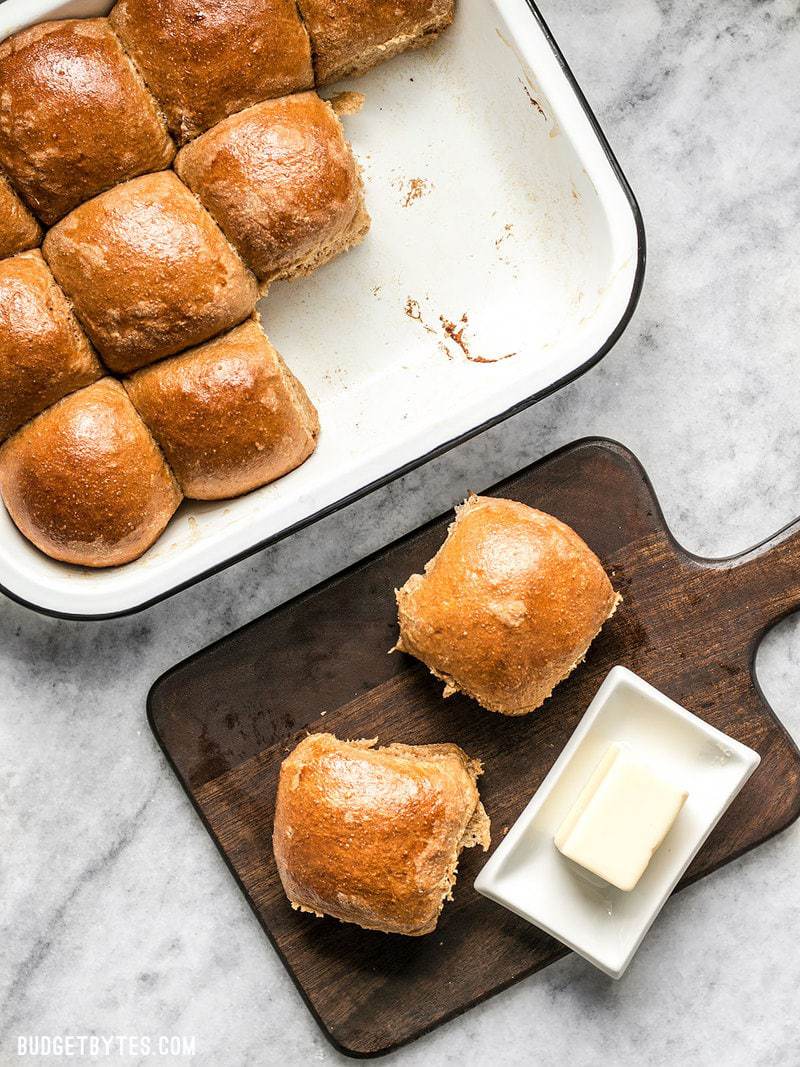 Two Sweet Molasses Dinner Rolls on a cutting board with butter next to the baking dish full of rolls