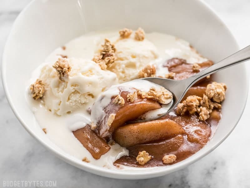 A bowl of Slow Cooker Hot Buttered Apples topped with ice cream and granola, being eaten with a spoon. 