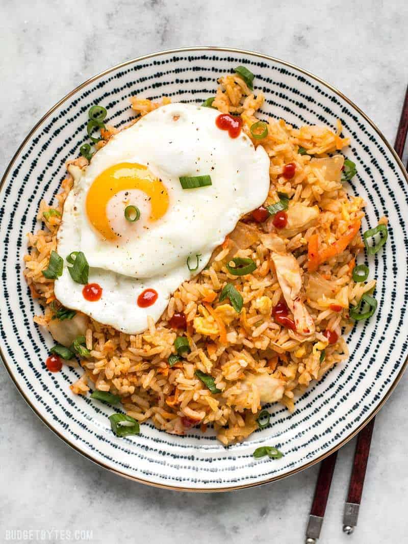 A plate full of Kimchi Fried topped with a fried egg, green onion, and sriracha