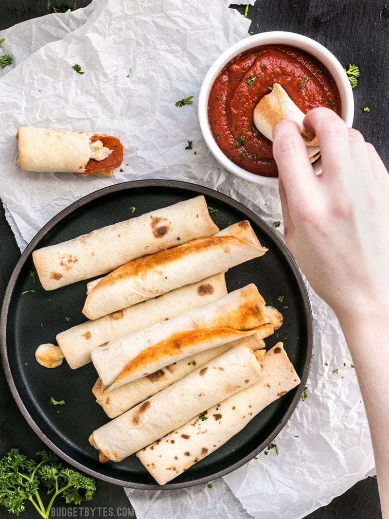A plate full of Pizza Roll Ups and a hand dipping one into a bowl of pizza sauce