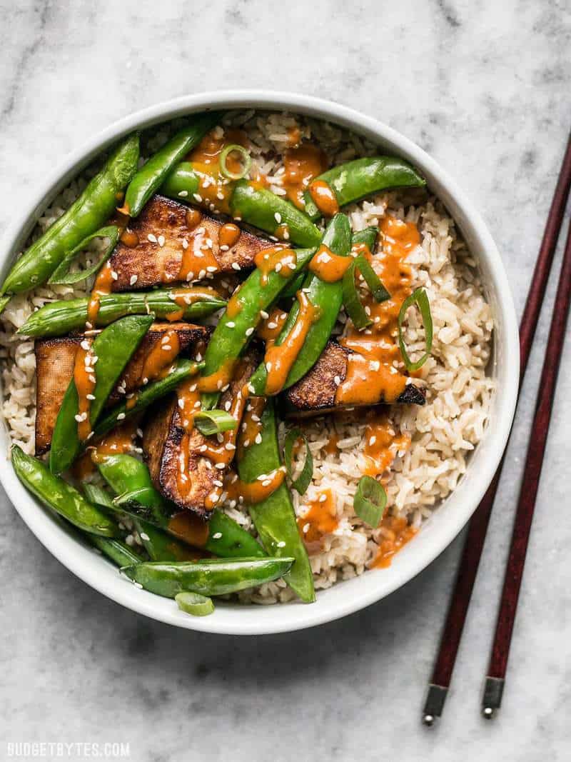 A bowl of Soy Marinated Tofu with brown rice, snow peas, and spicy peanut sauce drizzled over top