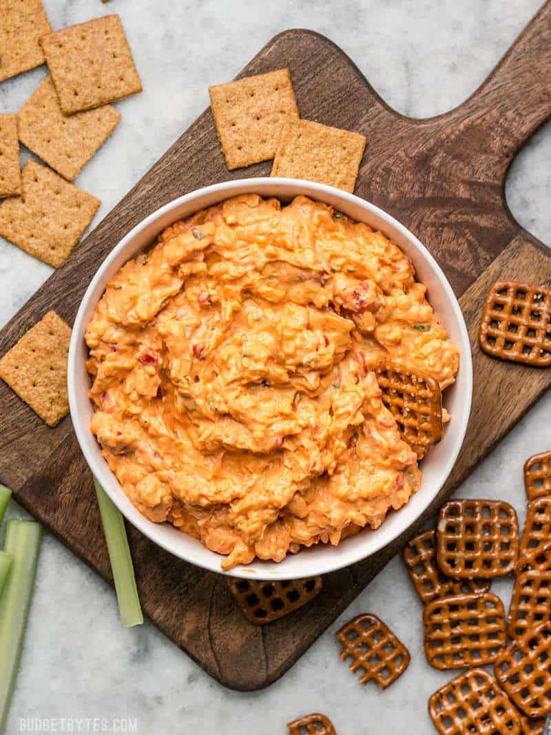 A bowl of Sriracha Pimento Cheese Spread on a wooden cutting board surrounded by crackers and pretzels