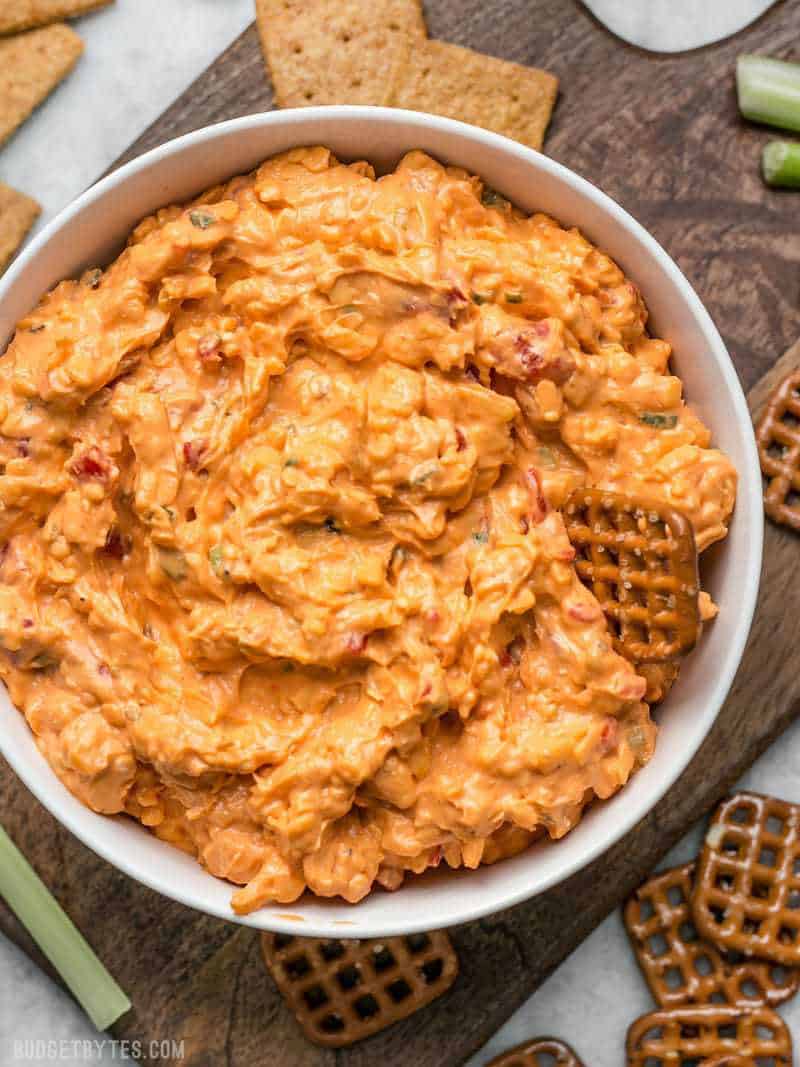 A bowl of Sriracha Pimento Cheese Spread surrounded by crackers, pretzels, and celery