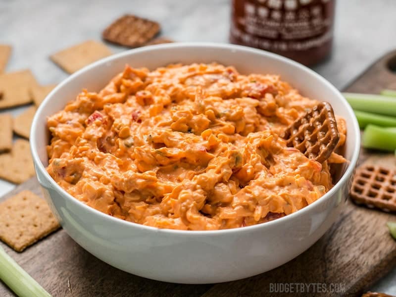 A pretzel being dipped into a bowl of Sriracha Pimento Cheese Spread 