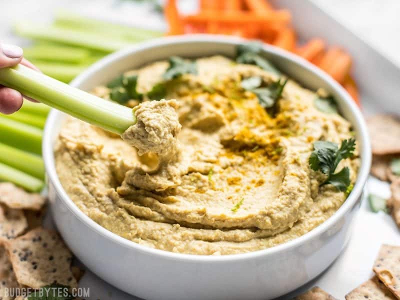 A celery stick being dipped into a bowl of Coconut Curry Hummus. 