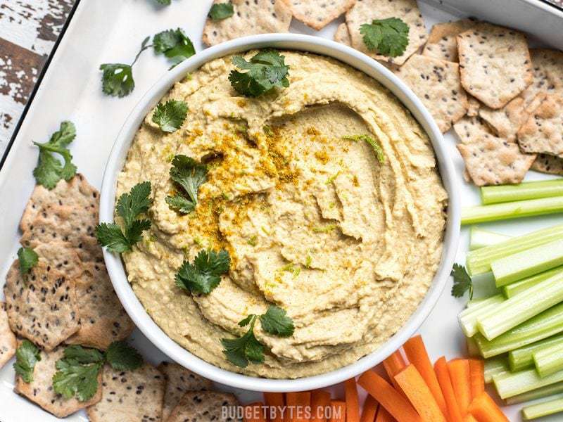 Cilantro sprinkled over a bowl of Coconut Curry Hummus surrounded by crackers and vegetable sticks 