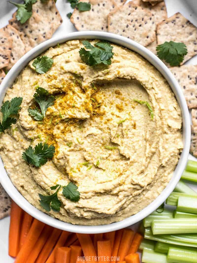 This Coconut Curry Hummus throws a new twist on the traditional dip with creamy coconut milk, vibrant lime, and warm curry spices. BudgetBytes.com A big bowl of Coconut Curry Hummus topped with cilantro and surrounded by vegetable sticks and crackers.