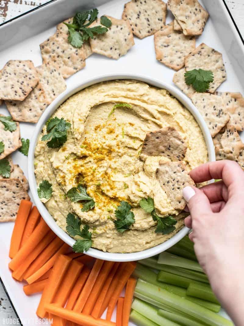 A platter of vegetables, crackers, and coconut curry hummus, and a hand dipping a cracker into the hummus. 