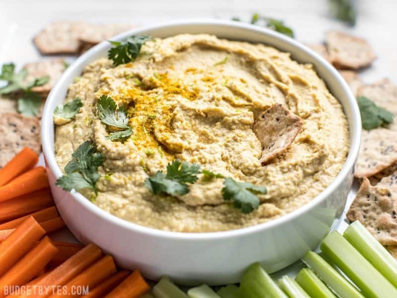 Front view of a bowl of Coconut Curry Hummus surrounded by vegetable sticks and crackers. 