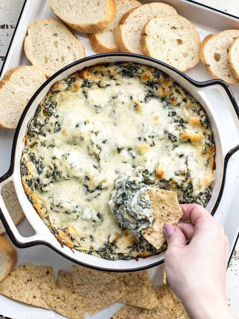 A hand scooping up some Double Spinach Artichoke Dip on a tortilla chip