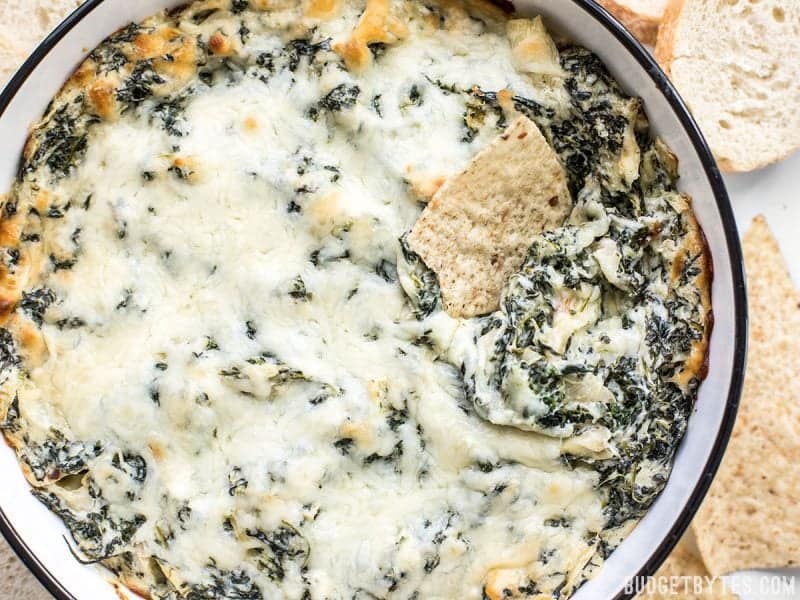 Close up of the casserole dish with Double Spinach Artichoke Dip surrounded by bread and tortilla chips