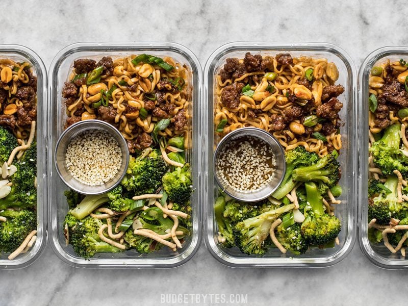 Four rectangular glass meal prep containers with Pork and Peanut Dragon Noodles, broccoli salad, and containers of salad dressing.