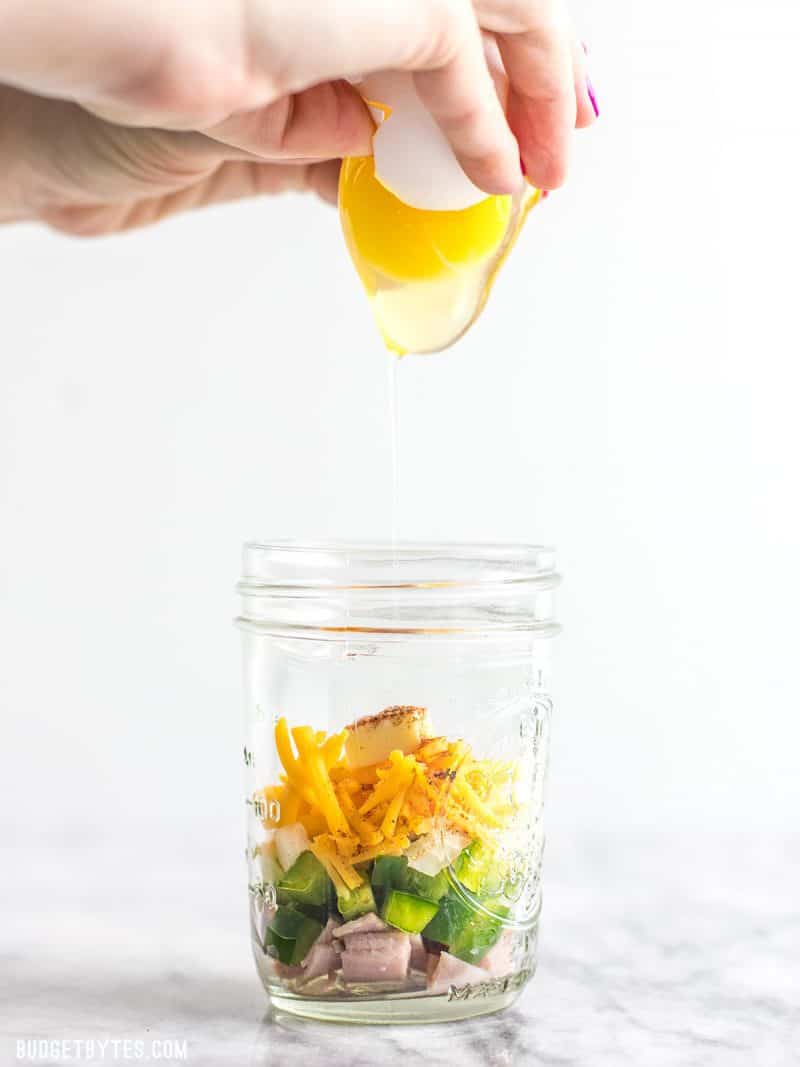 Make Ahead Microwave Breakfast Scrambles are a fast, easy, and healthy breakfast when you’re short on time! BudgetBytes.com An egg being cracked into a jar of Make Ahead Microwave Breakfast Scramble ingredients.