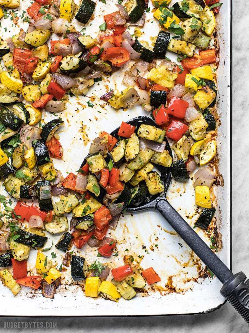 A white sheet pan full of roasted summer vegetables being scooped up by a spatula.