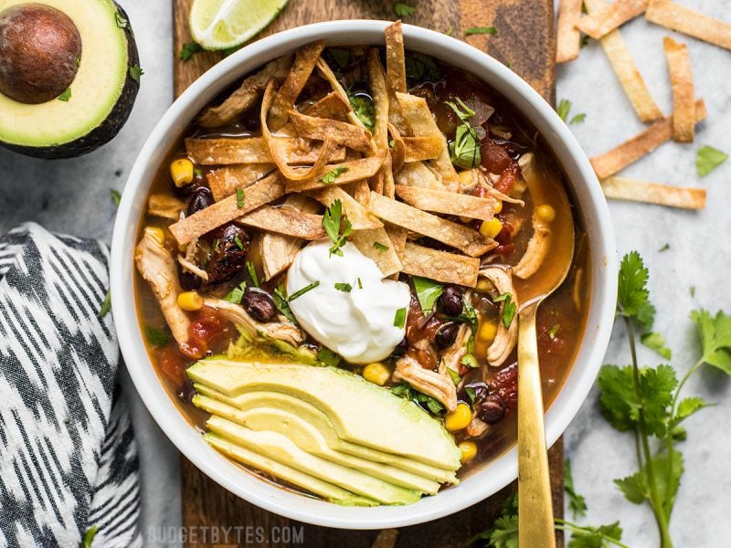 This Slow Cooker Chicken Tortilla Soup offers a deep, comforting, smoky flavor with simple ingredients and minimal effort. BudgetBytes.com Overhead view of a bowl of Slow Cooker Chicken Tortilla Soup with all the toppings and a gold spoon in the bowl.