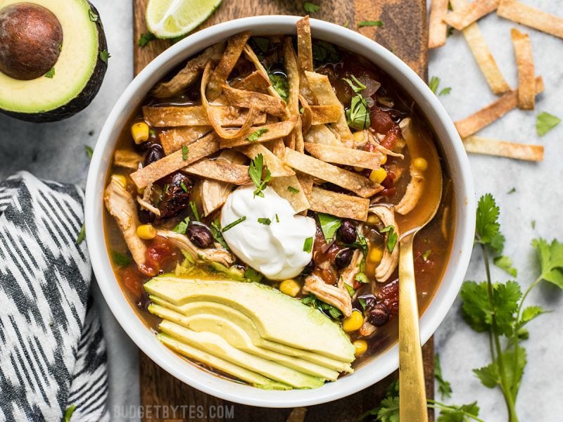 Overhead view of a bowl of Slow Cooker Chicken Tortilla Soup with all the toppings and a gold spoon in the bowl. 