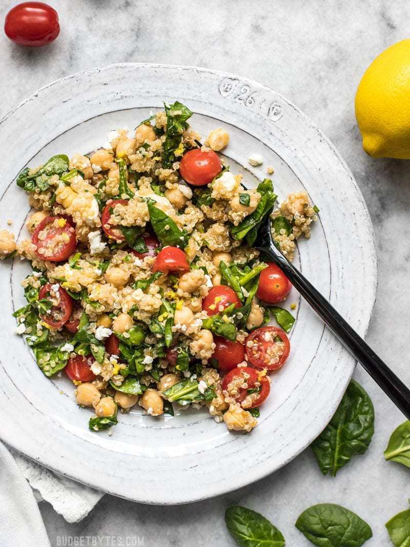 This Spinach Chickpea and Quinoa Salad is an awesome base to build meals throughout the week and it holds up extremely well in the fridge, so you can eat better with less effort. BudgetBytes.com A plate full of Spinach Chickpea and Quinoa Salad with spinach, tomatoes, and lemons scattered around
