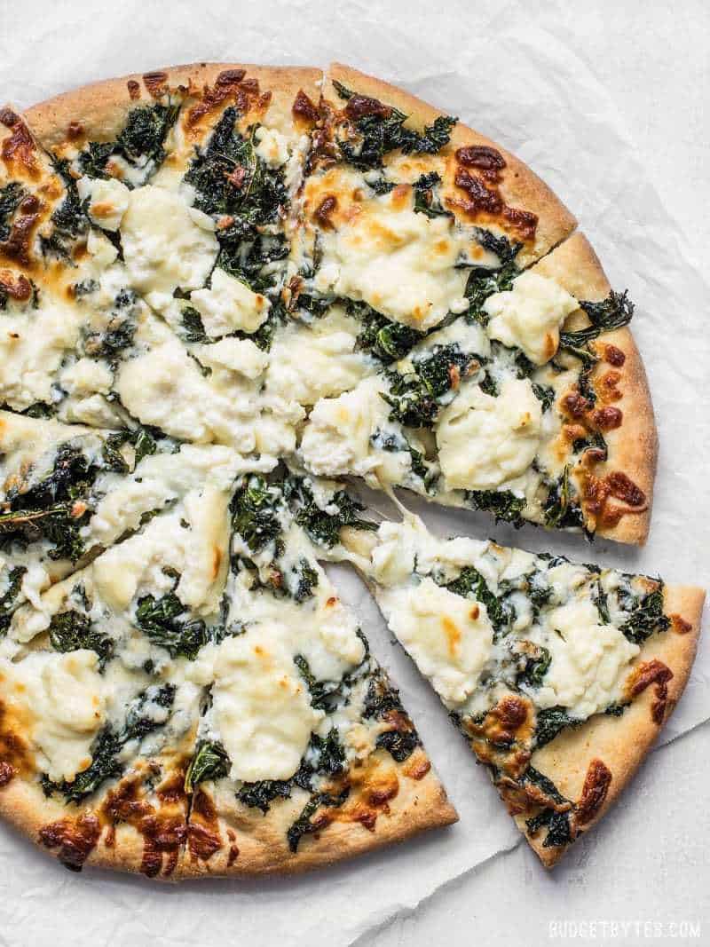 Garlicky greens and creamy ricotta pair perfectly on this light and fresh Garlicky Kale and Ricotta Pizza. It’s the perfect pizza for summer! BudgetBytes.com Baked Garlicky Kale and Ricotta Pizza on parchment with one slice being pulled away