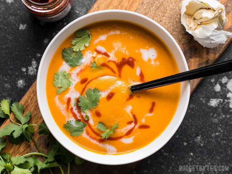 A spoon sinking into a bowl of Thai Coconut Curry Carrot Soup with coconut milk and sriracha swirled on top. 