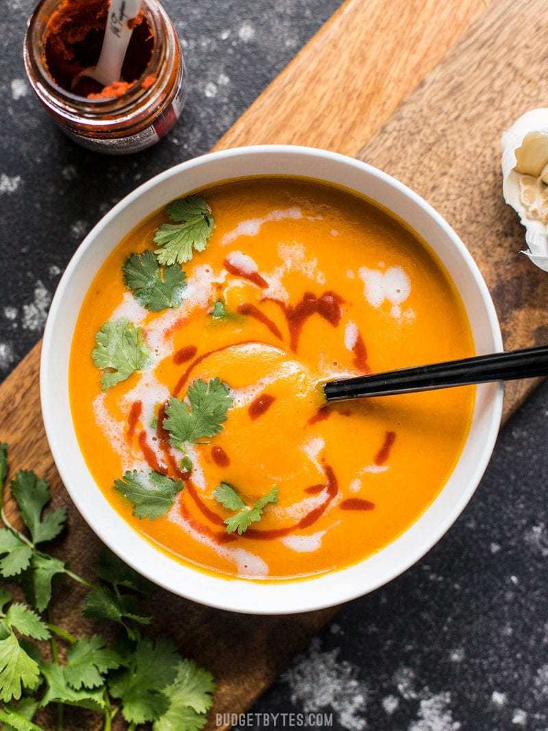 A bowl of Thai Coconut Curry Carrot Soup on a wooden cutting board with coconut milk and sriracha swirled into the top