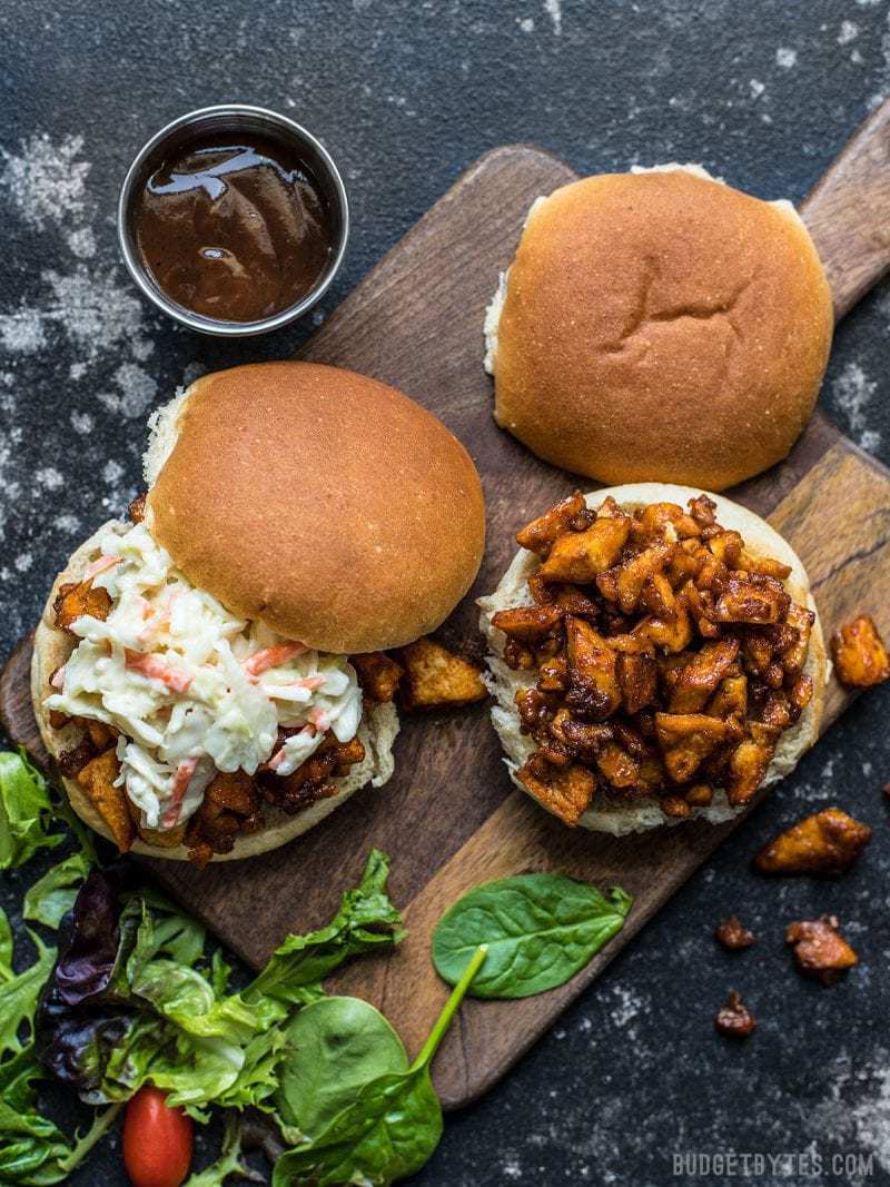 Two BBQ Tofu Sliders on a wooden cutting board with salad greens and a dish of BBQ sauce near by. 