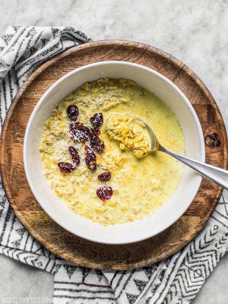 A bowl of Golden Milk Overnight Oats with dried cranberries and coconut, on a wood plate. 