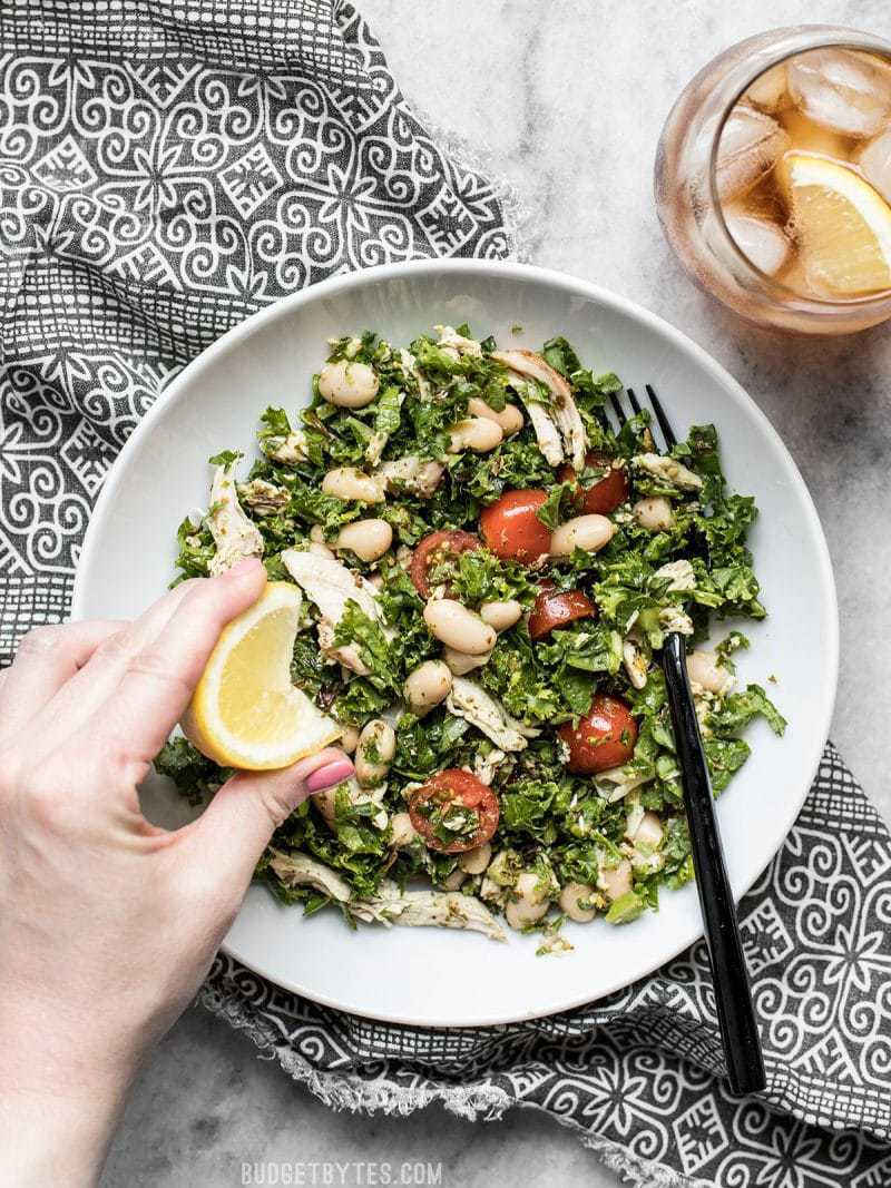 Lemon being squeezed onto a plate full of Kale White Bean and Pesto Salad with iced tea on the side