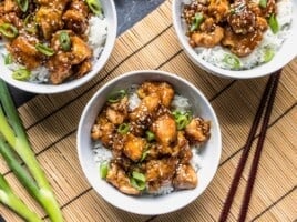 Overhead view of three bowls of sesame chicken with white rice and green onion.