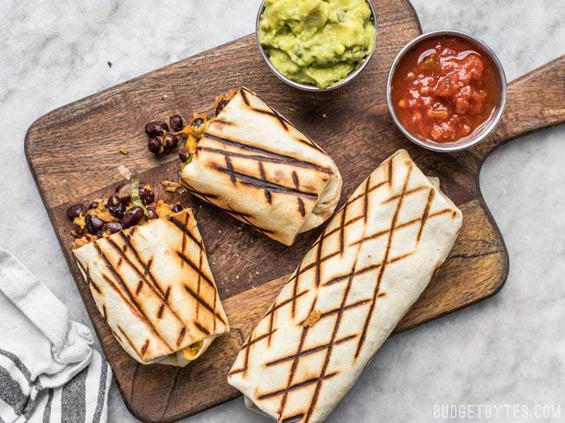 Two bean and cheese burritos on a cutting board, one cut in half, with cups of guacamole and salsa on the side. 