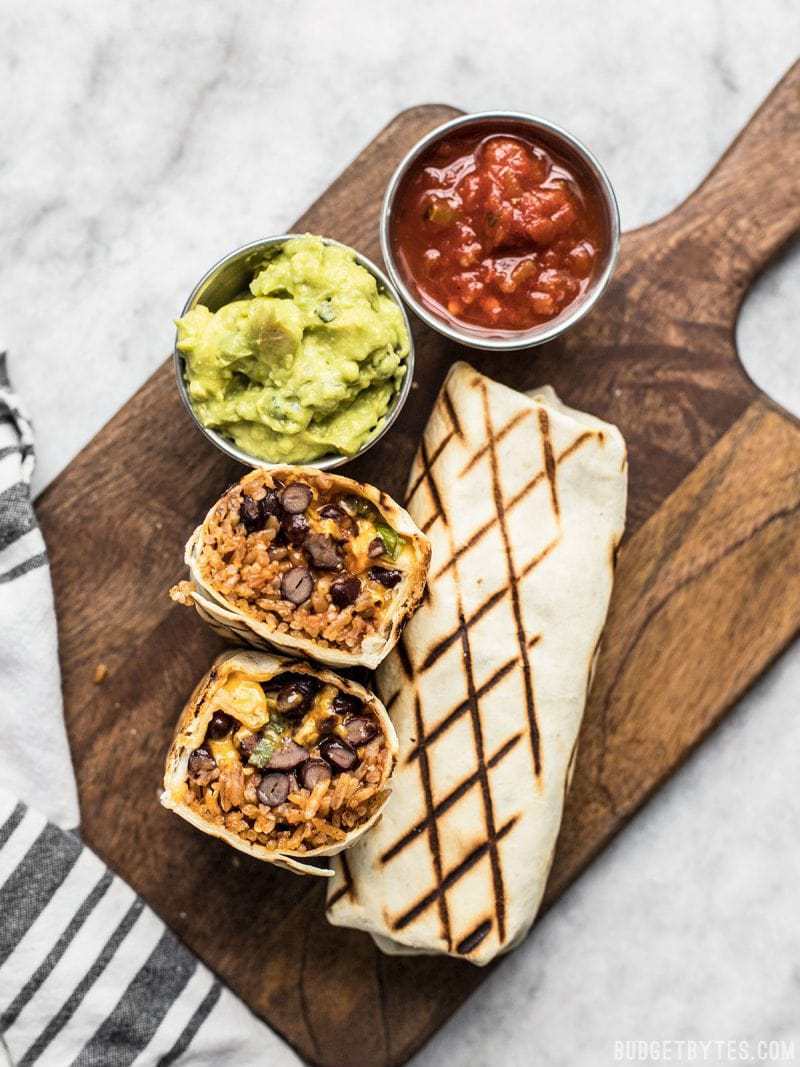 Two Bean and Cheese Burritos on a cutting board with cups of guacamole and salsa. 