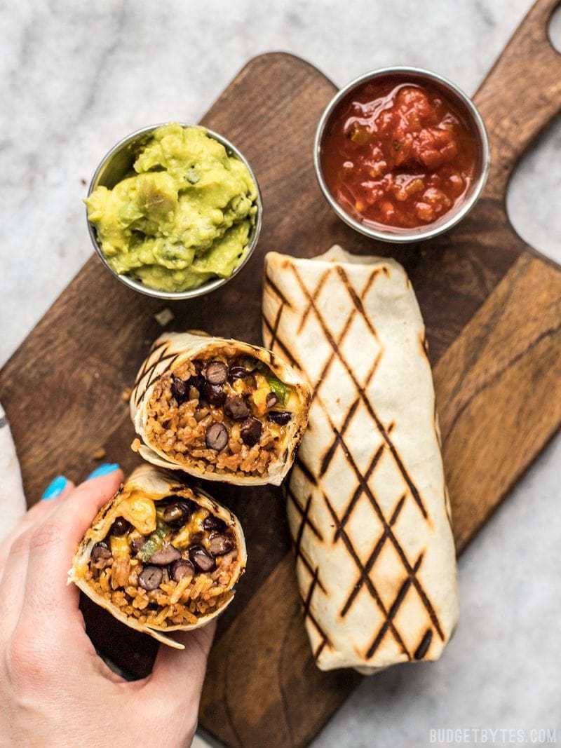 A hand holding a bean and cheese burrito ready to eat, with sides of guacamole and salsa.