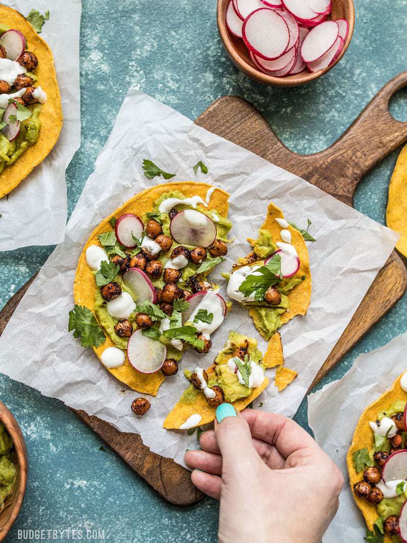 These cool and refreshing Spiced Chickpea Tostadas are the perfect almost no cook dinner for summer. Customize the toppings based on what you have on hand to make the most of leftovers! Budgetbytes.com A hand taking a piece of a Spiced Chickpea Tostada