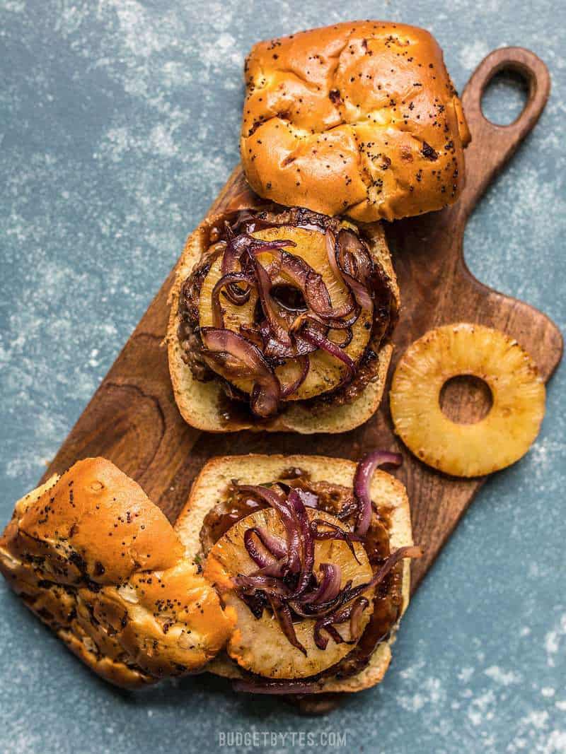 Two Hawaiian Burgers on a wooden cutting board with onion rolls and pineapple rings