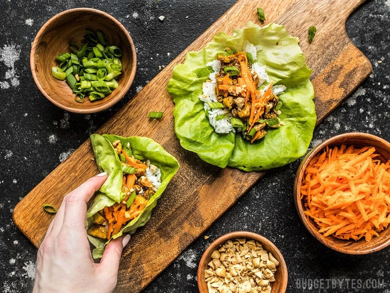 A hand taking one Hoisin Tofu Lettuce Cup from a wooden cutting board with toppings nearby