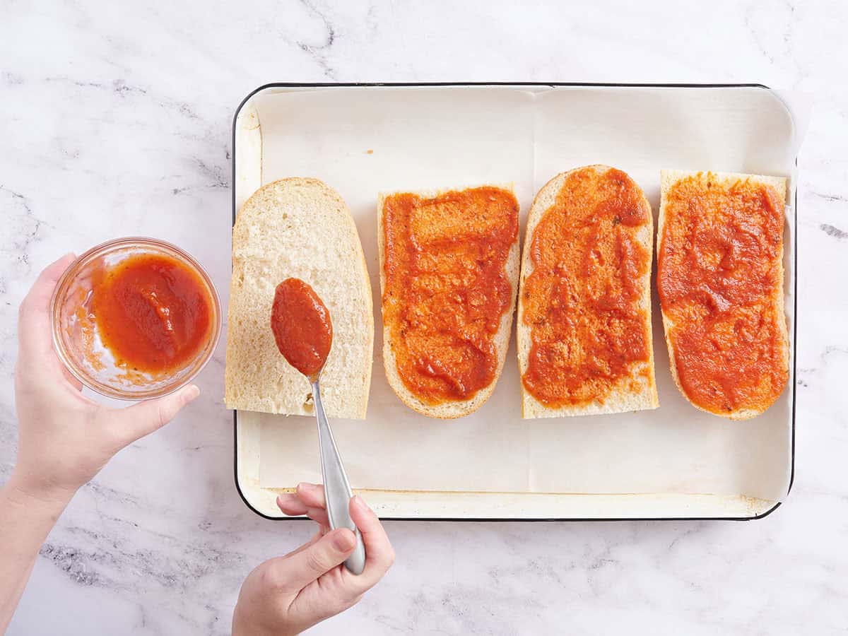 A hand spooning pizza sauce onto French bread quarters.