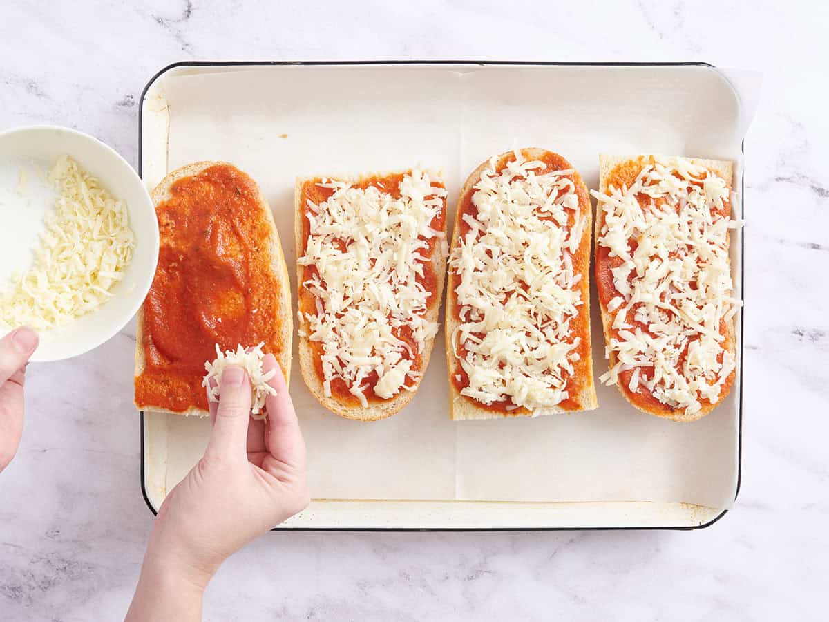 A hand sprinkling shredded cheese over French bread pizzas.
