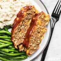 Two slices of Cheddar Cheeseburger Meatloaf on a dinner plate with mashed potatoes and green beans